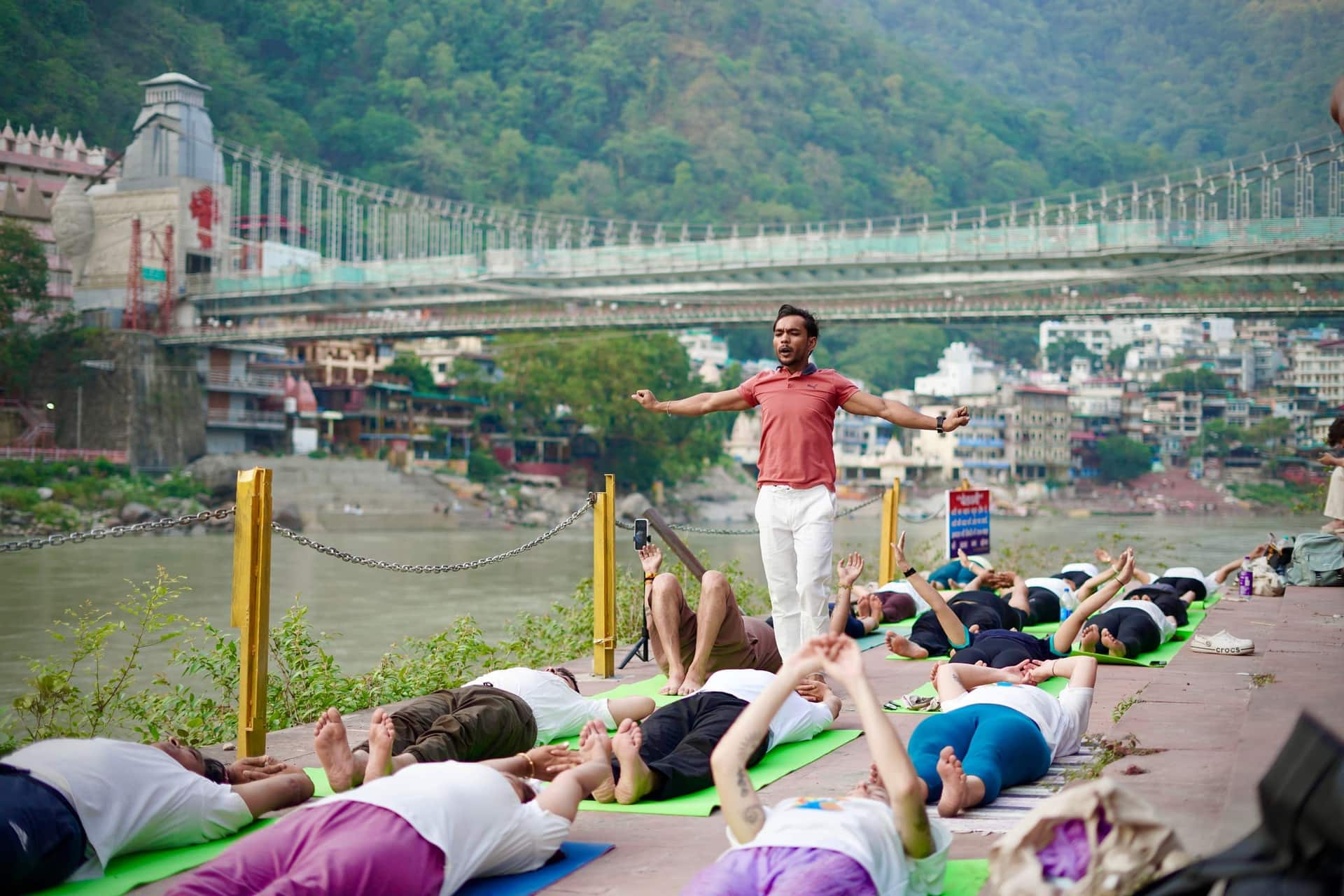 A yoga teacher guiding a small group through a posture on mats beside the river in Rishikesh