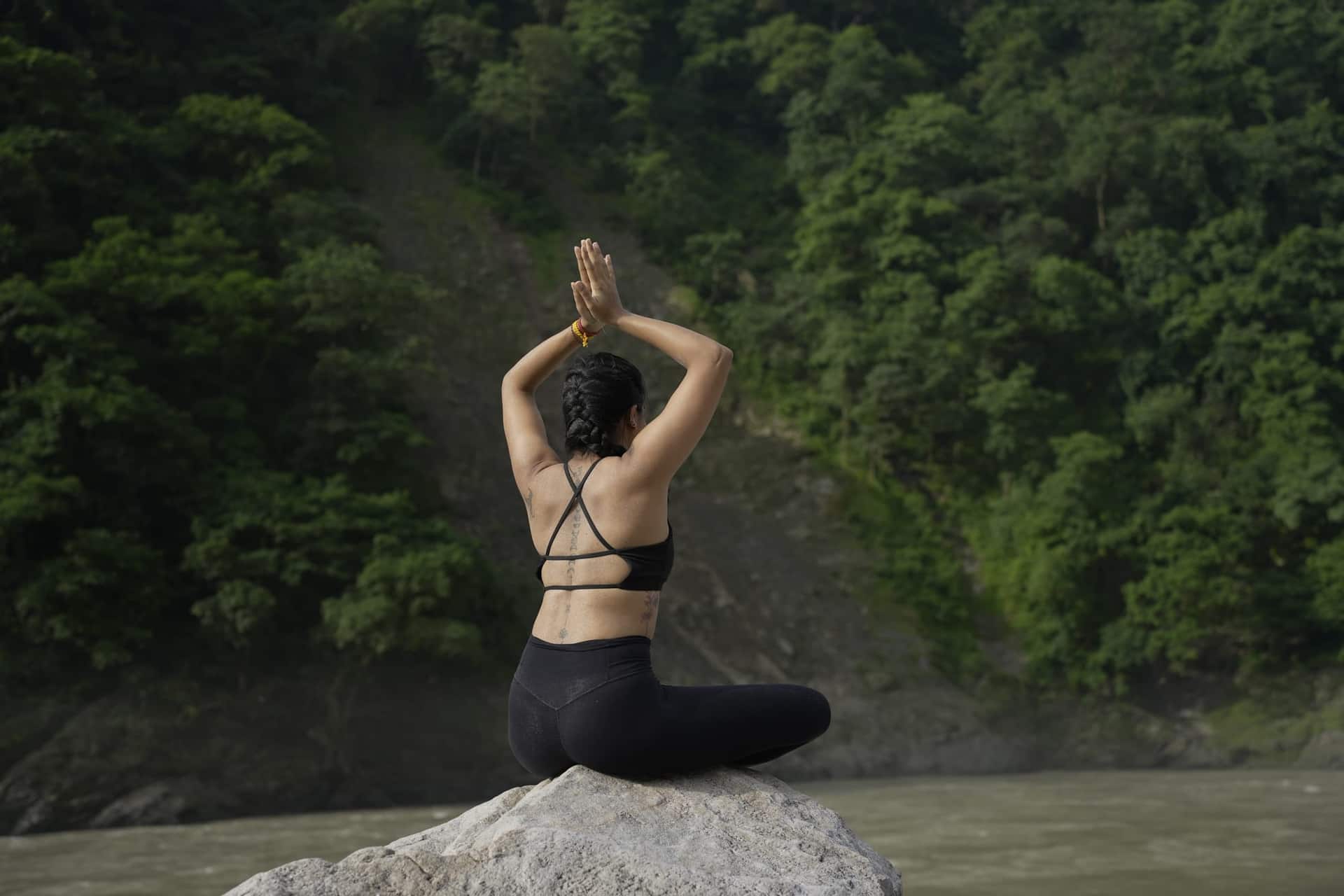 A woman sitting cross-legged on a boulder above the Ganga with her palms pressed together overhead