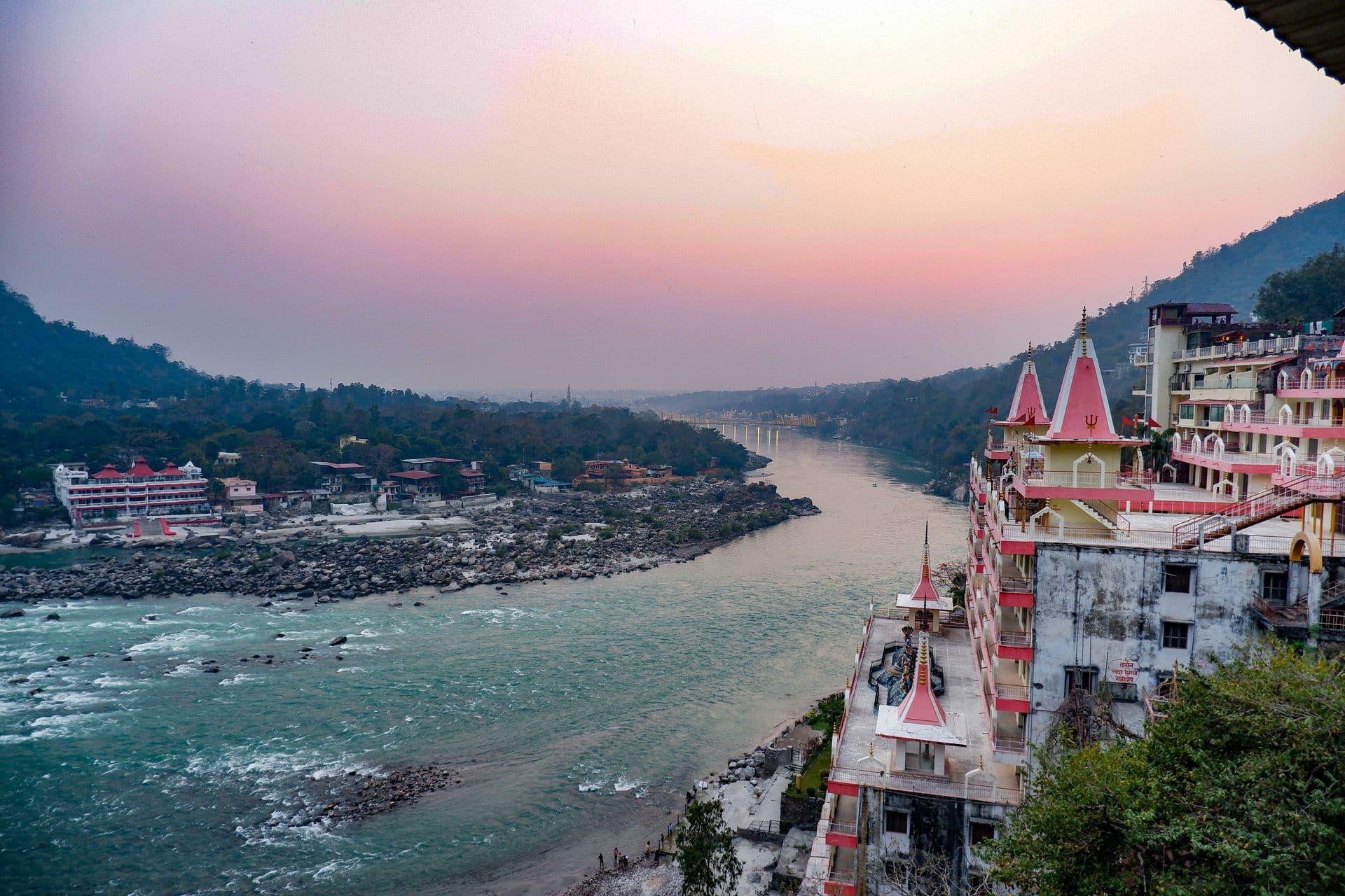Dusk over the turquoise Ganga at Rishikesh: the tiered pink Trayambakeshwar Temple on the right bank, the Laxman Jhula bridge faint downstream, forested hills and a peach sunset sky